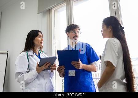 Ein medizinisches Team aus zwei Ärzten und einer Krankenschwester führt Gespräche an einem Fenster. In weißen und blauen Uniformen diskutieren sie die Patientenversorgung mit Tisch Stockfoto