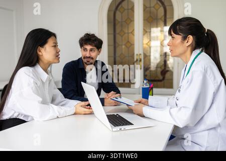 Ein Arzt in einem weißen Mantel bespricht die medizinische Versorgung mit zwei Patienten in einem hellen Büro. Sie führen aktiv Gespräche, mit einem Laptop und medizinischen Geräten Stockfoto