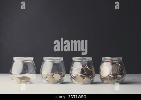Sammlung von Glasgläsern gefüllt mit Münzen, angeordnet in einer Reihe vor dunklem Hintergrund Stockfoto