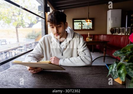 Junger Mann in lässigem Hoodie, der in einem gemütlichen Café mit Holztischen und großen Fenstern mit Blick auf die Außenterrasse eine Speisekarte liest. Sanfte Beleuchtung ergänzt den Stockfoto