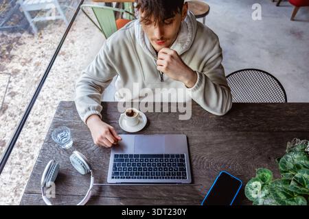 Ein junger Mann sitzt an einem rustikalen Tisch und arbeitet an seinem Laptop, umgeben von einer Tasse Kaffee, Kopfhörern und einem Smartphone, verkörpert den freischaffenden Tech li Stockfoto