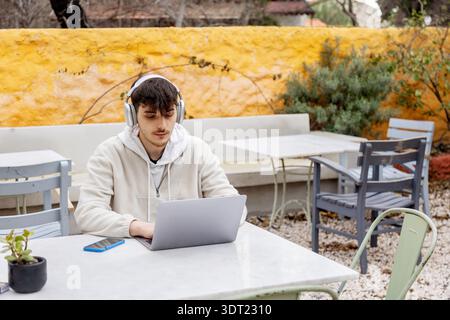 Ein junger Mann, der Kopfhörer trägt, arbeitet im Freien an einem Laptop, der freiberuflich arbeitet und Technologie verkörpert. Umgeben von der Natur konzentriert er sich auf seine technischen Aufgaben in Stockfoto