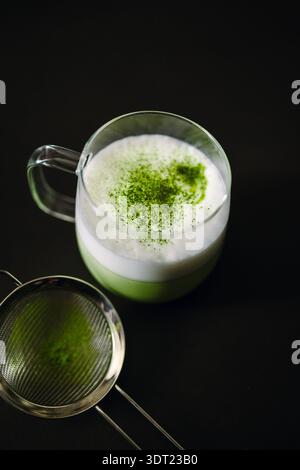 Ein cremiger Matcha-Latte in einer Tasse aus klarem Glas sitzt auf einer dunklen Oberfläche und zeigt Schichten von aufgeschäumter Milch und leuchtend grünem Matcha, mit einem Sieb in der Nähe, Ex Stockfoto