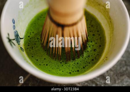 Nahaufnahme eines Bambus-Schneebesen, der leuchtend grünen Matcha in einer Keramikschale rührt. Der Mischprozess erzeugt eine glatte, schaumige Textur, die t hervorhebt Stockfoto