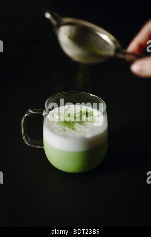 Ein frisch zubereiteter Matcha-Latte in einem klaren Glasbecher, der kräftigen grünen Tee unter einer üppigen Schaumschicht zeigt. Der dunkle Hintergrund verleiht Eleganz Stockfoto