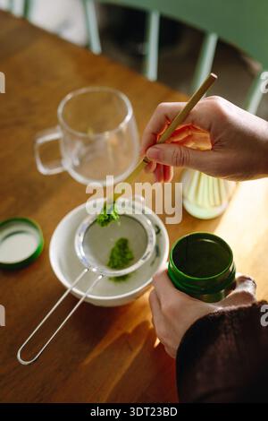 Eine Nahaufnahme einer Person, die sorgfältig Matcha-Tee zubereitete, mit einem Bambuslöffel das leuchtende grüne Pulver durch ein Sieb über einer weißen Schüssel siebt Stockfoto