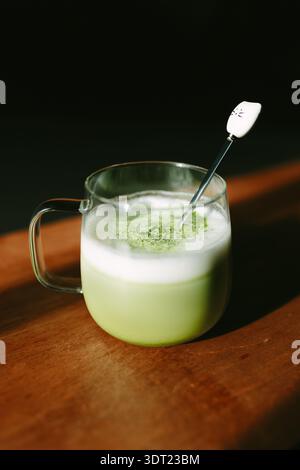 Eine warme Tasse Matcha Latte in einem klaren Glasbecher auf einer Holzoberfläche. Der aufschäumende grüne Tee wird mit Schaum belegt, der mit einem Löffel mit einem Kun gerührt wird Stockfoto