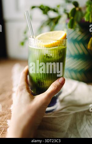 Eine Hand, die ein erfrischendes Glas Matcha und Lavendeleistee hält, garniert mit einer Zitronenscheibe. Das Getränk wird in einem gerippten Glas mit Strohhalm serviert Stockfoto