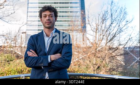 Ein selbstbewusster Geschäftsmann in blauem Anzug steht mit überkreuzten Armen im Freien vor einem Hintergrund von Bäumen und einem modernen Gebäude, das professionell ausstrahlt Stockfoto