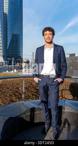 Ein junger professioneller Mann steht selbstbewusst in einem Business-Anzug draußen vor einer modernen Skyline der Stadt. Der klare blaue Himmel und die Architektur schaffen ein VI Stockfoto