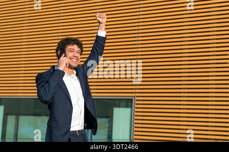 Geschäftsmann in Anzug feiert Erfolg, hebt seine Faust beim Telefonieren, steht draußen vor einer sonnigen, gestreiften Gebäudefassade und zeigt Stockfoto