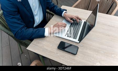 Geschäftsmann in blauem Anzug, der an einem Laptop auf einem Holztisch arbeitet, mit einem Handy neben ihm. Die Umgebung ist ein moderner Arbeitsbereich im Freien, fokussiert Stockfoto