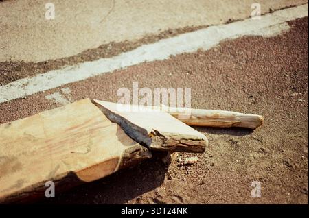 Gebrochene Holzdiele, die bei hellem Sonnenlicht auf einer Asphaltoberfläche liegt, minimale Zusammensetzung mit rauen Texturen und Schatten. Stockfoto