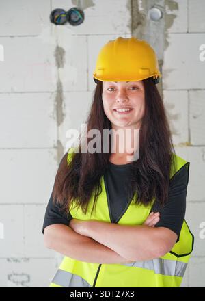 Junge Frau in gelbem Schutzhelm und grüner Warnweste, langes dunkles Haar, gekreuzte Hände, Blick in die Kamera. Verschwommene Baustellenwand hinten Stockfoto
