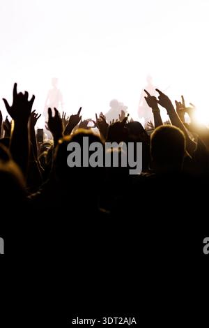 Ein Blickwinkel aus der Menge bei einem Musikfestival mit Silhouetten von Fans, die Rock-Schilder machen. Helle Bühnenlichter heben das Band während der cr-Funktion hervor Stockfoto