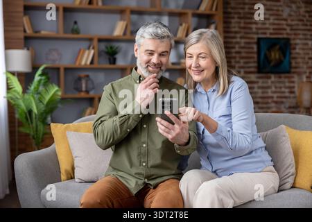 Lächelndes, reifes Paar genießt die gemeinsame Zeit, surft Inhalte auf einem Mobiltelefon und entspannt sich auf einem bequemen Sofa in ihrem modernen Wohnzimmer, das Verbindung und Technologie repräsentiert Stockfoto