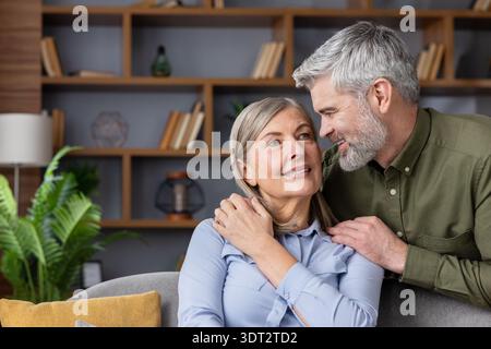 Ein älterer Ehemann nimmt seine lächelnde Frau sanft in ihr gemütliches Wohnzimmer ein und teilt einen warmen, liebevollen Moment, der dauerhafte Liebe, Vertrauen und komfortable Gesellschaft widerspiegelt Stockfoto