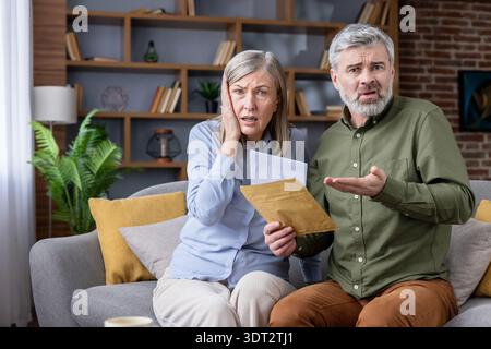 Unglückliches Seniorenpaar, das zu Hause auf dem Sofa sitzt und einen offiziellen Brief mit schlechten Nachrichten erhält, der Schock und Verwirrung über finanzielle Probleme und Schulden zeigt Stockfoto