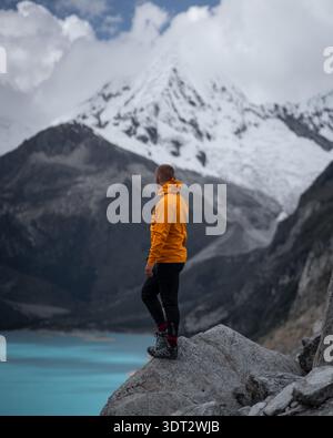 Blick auf einen Mann in einer gelben Jacke auf einem Felsen mit Blick auf den türkisfarbenen See und die schneebedeckten Berge, Ancash, Peru. Stockfoto