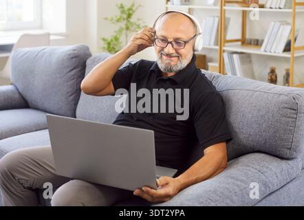 Lächelnder älterer Mann mit Kopfhörern, der während der Online-Arbeit einen Laptop auf dem Sofa benutzt Stockfoto