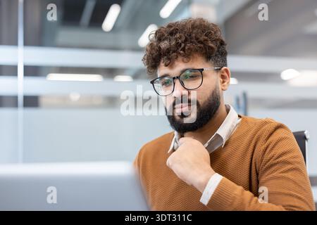 Junger männlicher Angestellter in Brille und braunem Pullover, konzentriert, arbeitet an einem Laptop in einem modernen Büro, repräsentiert Konzepte des Geschäfts, Innovation Stockfoto