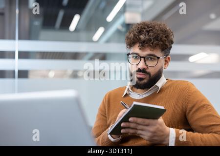 Junger Mann mit lockigem Haar und Bart, der eine Brille trägt, konzentrierte sich auf den Laptop, während er in einem modernen Coworking-Büro Notizen in ein Notizbuch schreibt, plant und rekonstruiert Stockfoto
