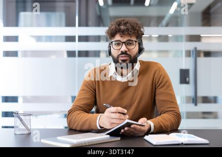 Junger Mann mit Headset und Brille, aktiver Teilnahme an einer Videokonferenz von seinem Schreibtisch aus, Blick auf den Zuschauer und Notizen Stockfoto