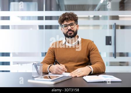 Junger Geschäftsmann mit lässiger Kleidung und Brille, der am Schreibtisch sitzt, einen Stift hält und in einem Notizbuch schreibt, sich auf Papierkram und Stockfoto