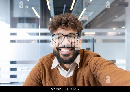 Junger professioneller Mann mit Bart und lockigem Haar, der eine Brille trägt und hell lächelt, während er einen Videoanruf macht oder ein Selfie in einem zeitgenössischen Offi macht Stockfoto