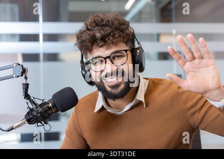 Junger Mann lächelt und winkt, während er Kopfhörer trägt und in ein Mikrofon spricht, live aus einem modernen Studio sendet und digitales Audio erzeugt Stockfoto