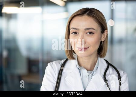 Junge Ärztin mit freundlichem Ausdruck lächelt in die Kamera, trägt ein medizinisches Peelinghemd, weißen Labormantel und Stethoskop, verkörpert Professionalität und Vertrauen im Gesundheitswesen Stockfoto