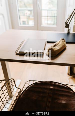 Minimaler Arbeitsbereich mit Notebook- und Bleistiftetui auf einem Holzschreibtisch in der Nähe eines hellen Fensters, mit weichem Tageslicht in einer modernen Einrichtung Stockfoto