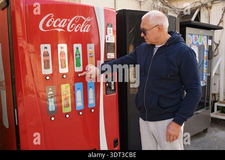 Gzira, Malta - 17. Februar 2026: Ein erwachsener Mann in einem blauen Hoodie, der ein Getränk aus einem roten Coca-Cola-Automaten wählt. Stockfoto