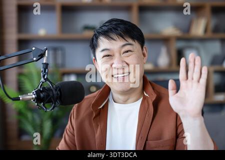 Asiatischer Mann, der Inhalte erstellt, lächelt und winkt, einen Livestream ausstrahlt oder einen Podcast in einem Heimstudio aufnimmt und sich mit seinem Online-Publikum verbindet Stockfoto