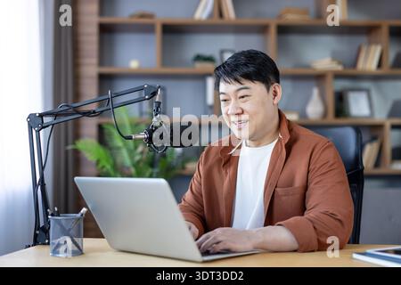 Asiatischer Mann, der an einem Schreibtisch sitzt und eine Online-Podcast-Episode aufnimmt, aktiv mit seinem Laptop und seinem professionellen Mikrofon in einem Heimstudio interagiert und digitale Inhalte für sein Publikum erstellt Stockfoto