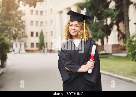 Der Absolvent steht stolz in Cap and Gown auf dem Campus, nachdem er während der Abschlussfeier das Diplom erhalten hat Stockfoto