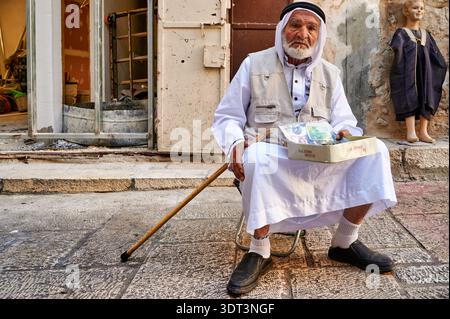 Ein alter Mann sitzt auf einem kleinen Stuhl in Jerusalems Altstadt. Er hält eine Kiste und sucht Hilfe von Passanten. Stockfoto