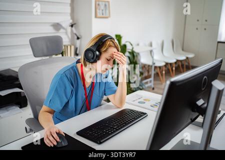Müde Krankenschwester mit Kopfhörern, die im Büro am Computer arbeiten Stockfoto