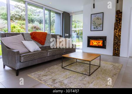 Dunkelgraues Ledersofa auf der linken Seite des modernen Wohnzimmers mit orangefarbenem Überwurf und Kamin Stockfoto