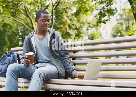 Mann sitzt tagsüber auf der Bank mit Laptop und Kaffee im Park Stockfoto