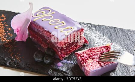 Moderne OPERA-Tortenscheibe mit leuchtender lila Glasur, zarter weißer Schokoladenkalligrafie und frischen Blaubeeren, ein vielseitiges Dessert, das r Stockfoto