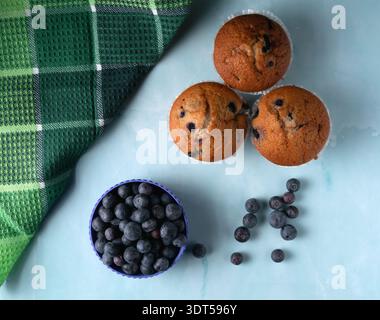 Blick von oben auf drei goldene Heidelbeermuffins, serviert mit einer Schüssel frischer Bio-Heidelbeeren auf einem hellen ästhetischen Hintergrund Stockfoto