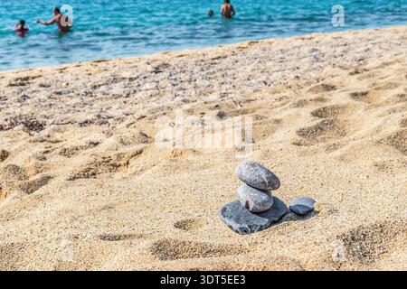Horizontale Aufnahme einer kleinen Zen-Steinpyramide an einem Sandstrand mit einem verschwommenen Hintergrund von Menschen, die im türkisfarbenen Meer schwimmen Stockfoto
