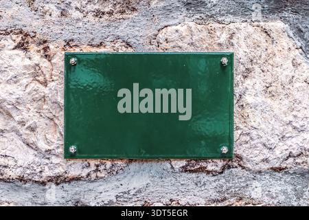 Leere grüne Metallplatte, die mit Schrauben an einer strukturierten Steinwand befestigt ist. Leeres Vorzeigemodell auf altem Steinhintergrund für benutzerdefinierten Text oder Hausnummern Stockfoto