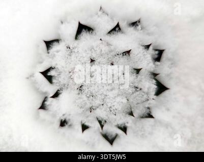 Runder Metallabfluss teilweise mit Neuschnee bedeckt. Kreisförmiges Muster mit dreieckigen Löchern, sichtbar durch weiße Schneeschicht, Ansicht von oben in Nahaufnahme Stockfoto