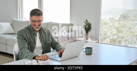 Ein glücklicher älterer Mann mittleren Alters, der einen Laptop am Heimtisch benutzt. Stockfoto