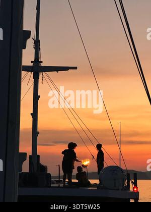 Segelszene bei Sonnenuntergang mit Silhouetten, zwei Personen an Deck und einem leuchtend orangefarbenen Himmel über dem Meer Stockfoto