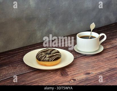 Eine Tasse schwarzen Kaffee und ein frischer Donut mit Schokoladenüberzug auf einem runden Teller auf einem Holztisch. Nahaufnahme. Stockfoto