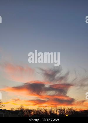 Amelia Ohio: ​A vertikale Niedrigwinkelaufnahme eines dramatischen Sonnenuntergangshimmels mit strukturierten, windgepeitschten Zirruswolken. Die Wolken sind in lebhaften Tönen von feurigem Orange und zartem Rosa gemalt und wirken wie Rauch oder Pinselstriche gegen einen Farbverlauf von blassem Blau bis tiefem Indigo. Darunter bilden die dunklen, blattlosen Silhouetten von Winterbäumen und ein Vorstadtdach den unteren Teil der Szene. Stockfoto
