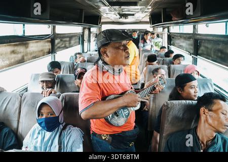 Ein Straßenmusiker unterhält die Passagiere in einem überfüllten öffentlichen Bus während einer täglichen Pendlerfahrt in Indonesien und steht mit seiner kleinen Gitarre im Gang Stockfoto